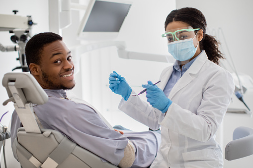Professional Teeth Cleaning. Black Female Dentist Having Treatment With Male Patient, Professional Stomatologist Woman In Medical Mask And Face Shield Holding Dental Tools And Looking At Camera