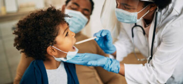 Family doctor examining throat of a small black boy while visiting him at home during coronavirus pandemic.